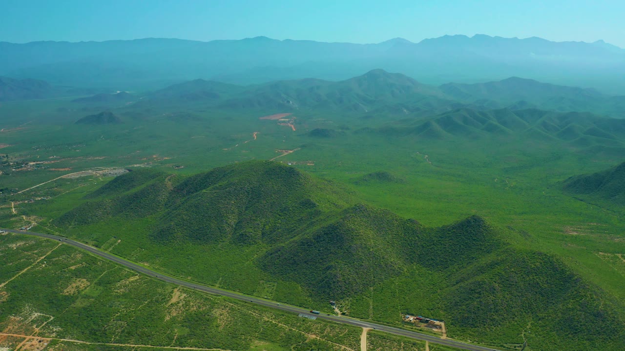 Drone-height view over Cerritos Beach inland terrain, showing a paved road at the bottom, green hills, pale dirt tracks, hazy mountain layers, and no visible people.