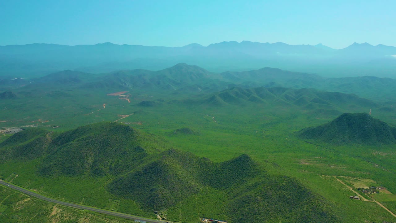 Aerial opening frame over green hills near Cerritos Beach, Baja California Sur, with a paved road in the lower foreground, distant mountain ridges, and no visible people.
