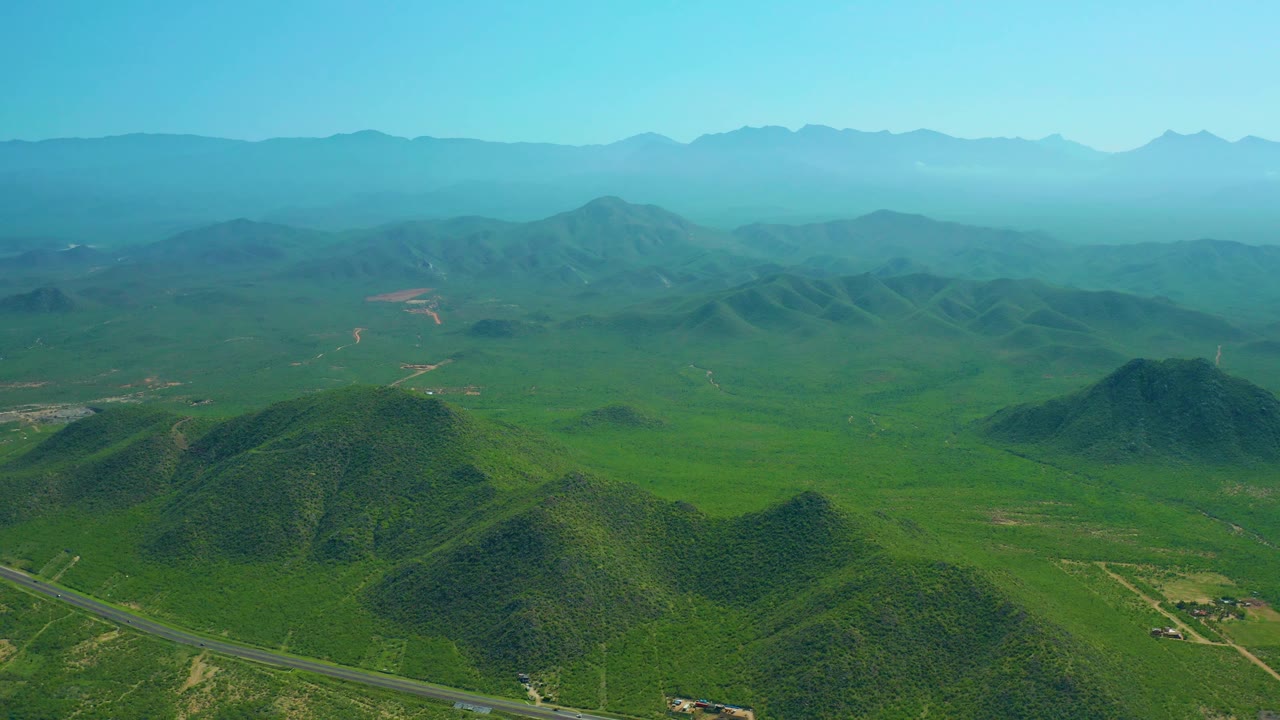 Wide aerial frame from Cerritos Beach, Baja California Sur, showing layered green hills, a road in the foreground, broad valley terrain, hazy distant mountains, and no visible people.