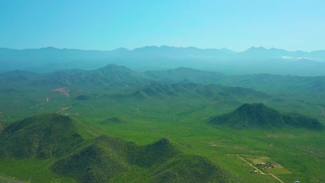 Wide aerial view over Cerritos Beach, Baja California Sur countryside, with green rounded hills in front, a broad valley, faint roads, hazy mountain layers, and no visible people.