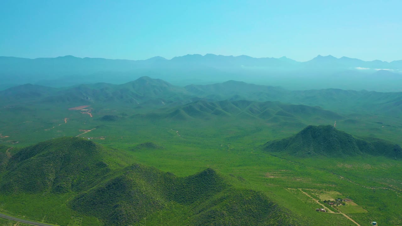 Cerritos Beach, Baja California Sur aerial view with a green valley floor, small road traces, clustered hills, distant mountains softened by haze, and no visible people.