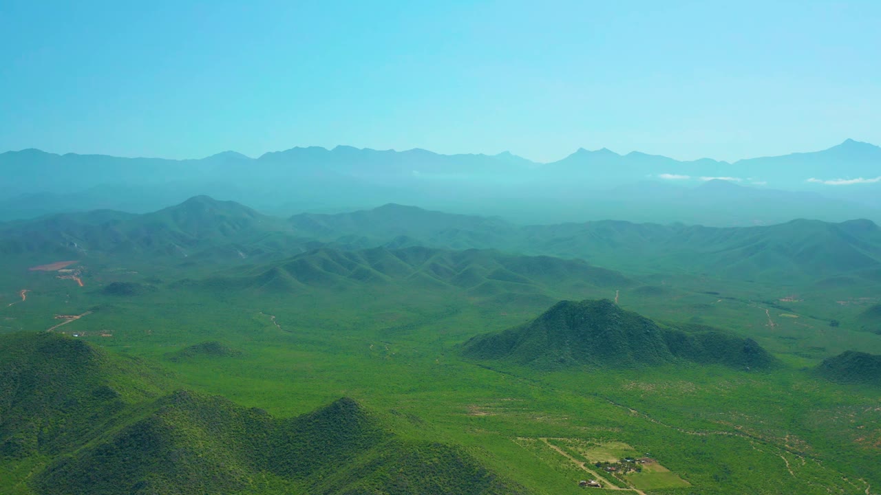 High aerial view near Cerritos Beach with rounded green hills in the foreground, a wide valley beyond, light haze, and no visible people.