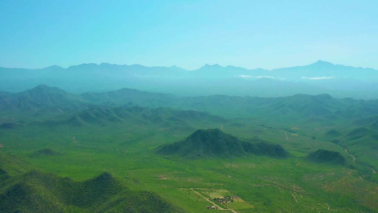 Wide aerial frame over green hills and open terrain near Cerritos Beach, Baja California Sur, with hazy mountains on the horizon and no visible people.