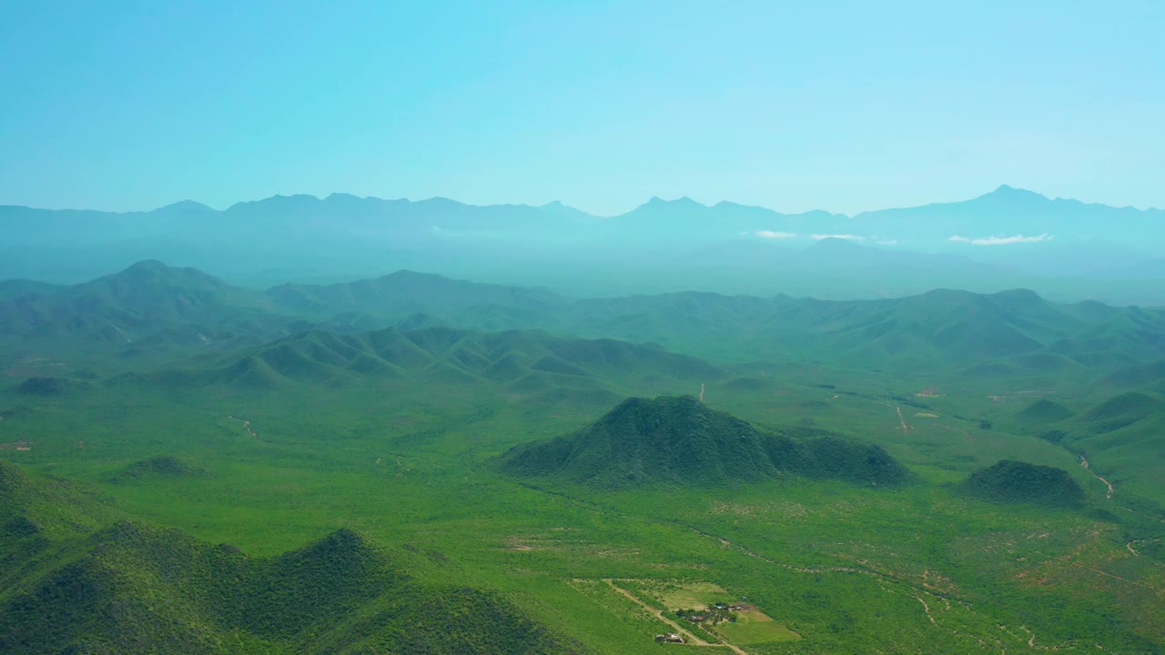 Wide drone frame near Cerritos Beach with green rolling terrain, scattered low hills, a distant mountain backdrop, light haze, and no visible people.