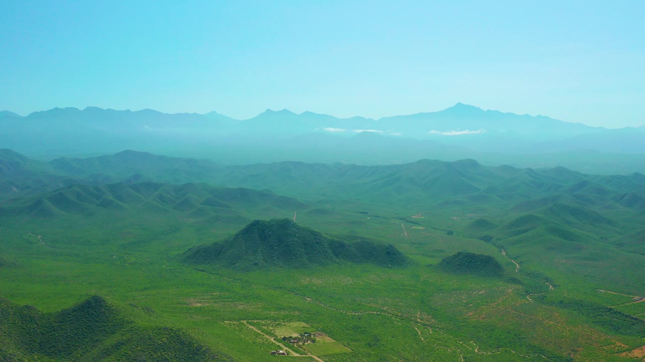 At 0:00, an aerial view over Cerritos Beach, Baja California Sur shows green foreground hills, wide valleys, hazy distant ridges, clear blue sky, and no visible people.