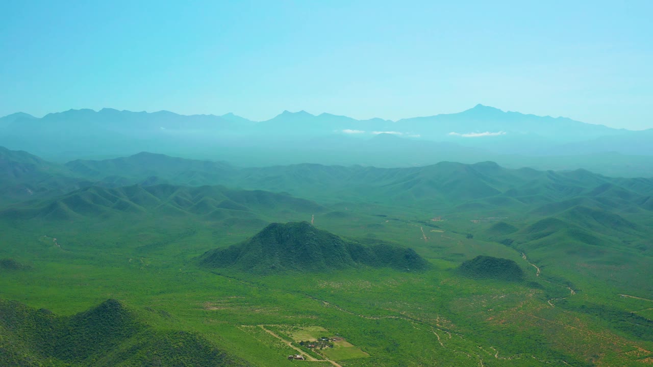 At 0:02, Cerritos Beach, Baja California Sur appears as green inland terrain with a small cluster of structures near the lower edge, low hills, distant peaks, clear sky, and no visible people.