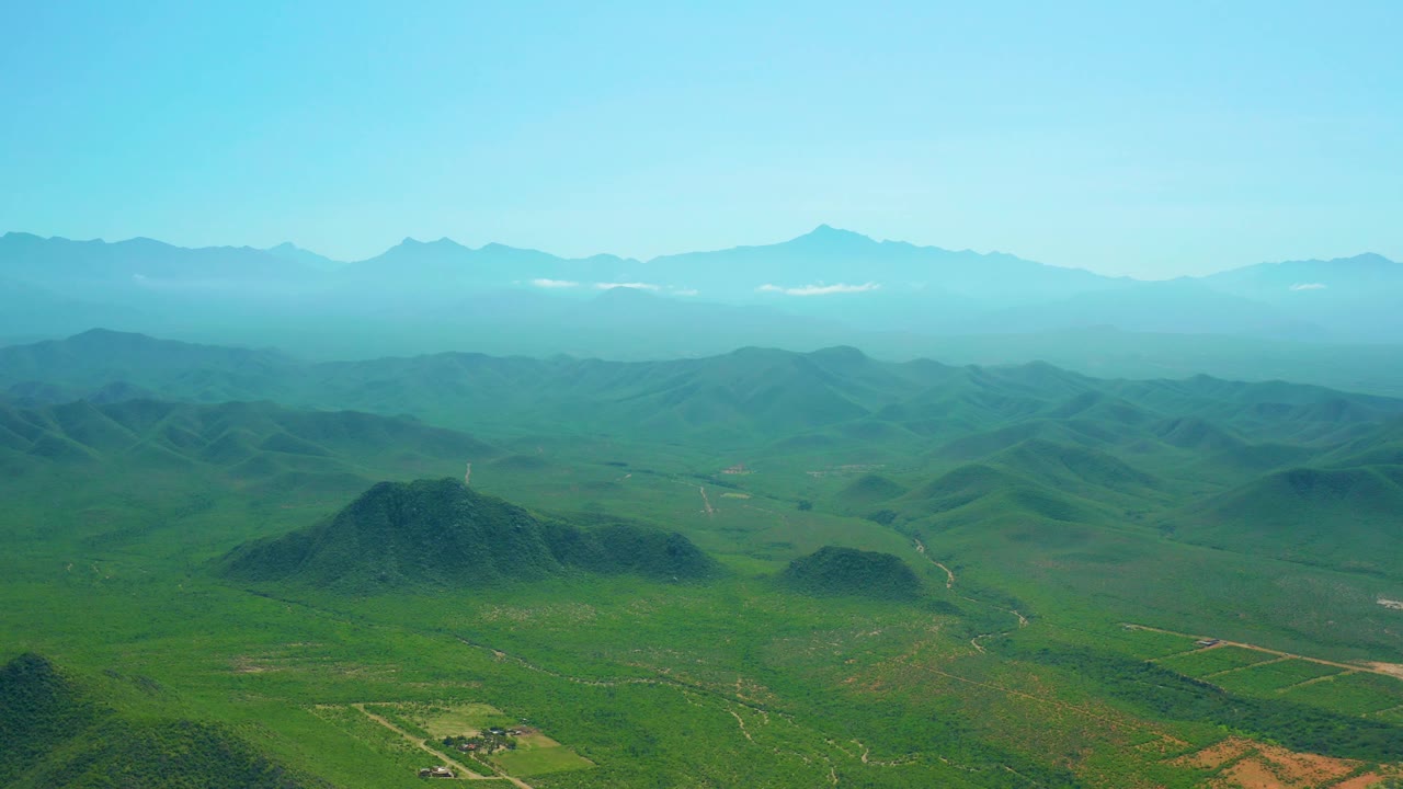 Cerritos Beach area aerial still with a green valley, low hills, faint dirt roads, small rural buildings near the lower edge, and no visible people.