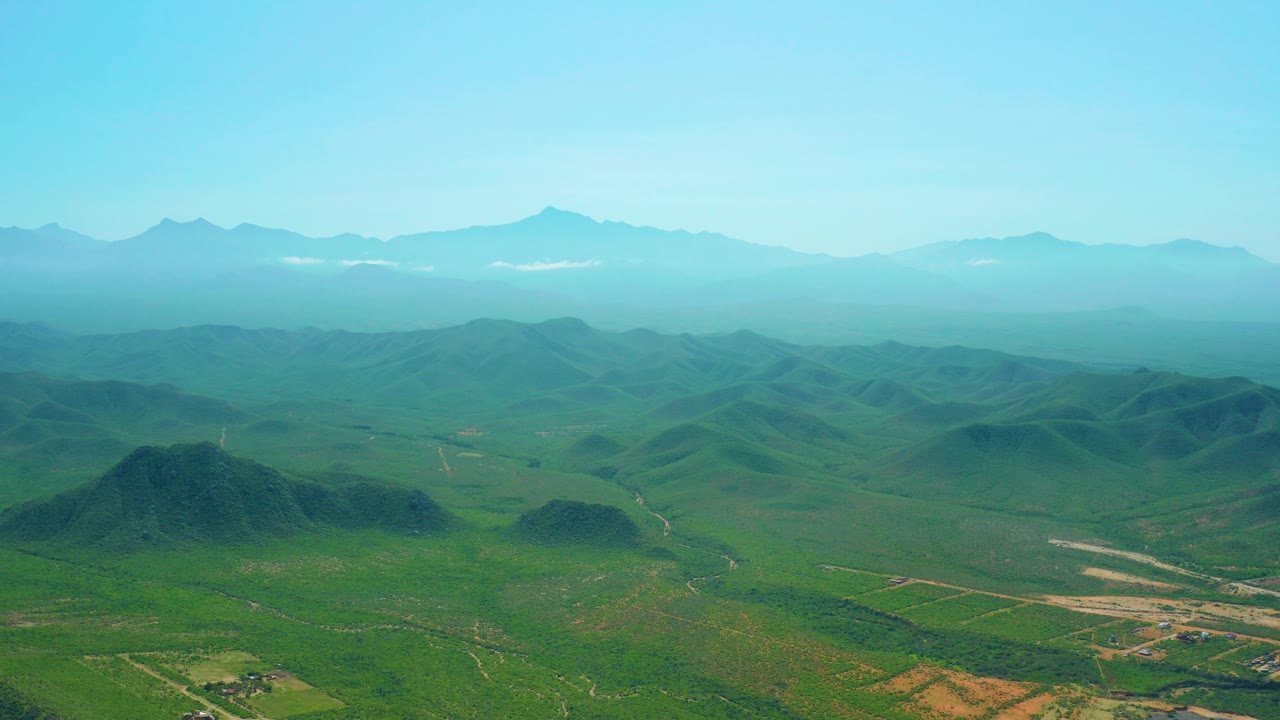 Wide aerial view of green hills and distant mountains under blue sky.