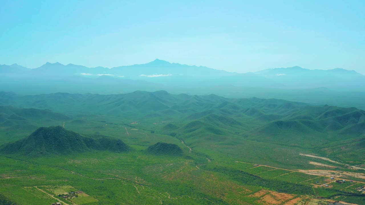 Aerial frame over Cerritos Beach inland terrain with rolling green hills, a pale road crossing the valley, blue mountain silhouettes, and no visible people.