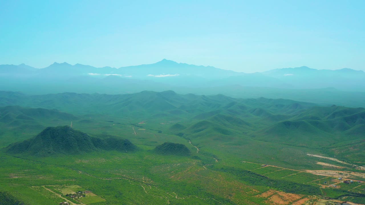 Drone-height view near Cerritos Beach showing low green hills, branching dirt roads, a broad hazy horizon, distant mountains, and no visible people.
