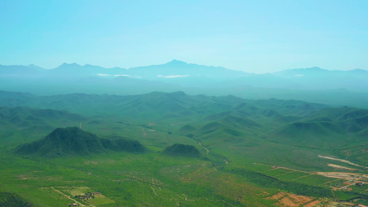 Wide Cerritos Beach area landscape with a rounded green hill in the lower frame, dirt tracks across the valley, layered blue mountains, and no visible people.