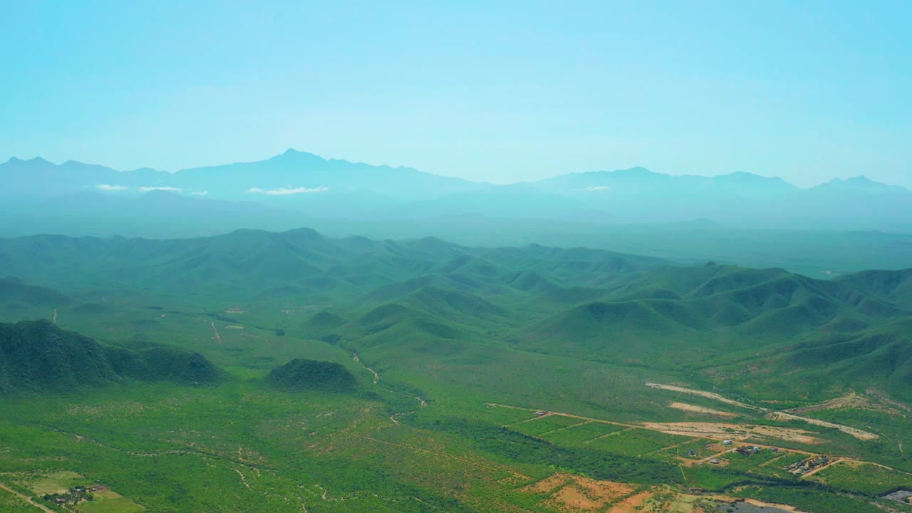 Aerial view near Cerritos Beach showing layered green hills, a pale road network in the lower valley, hazy mountain peaks on the horizon, and no visible people.