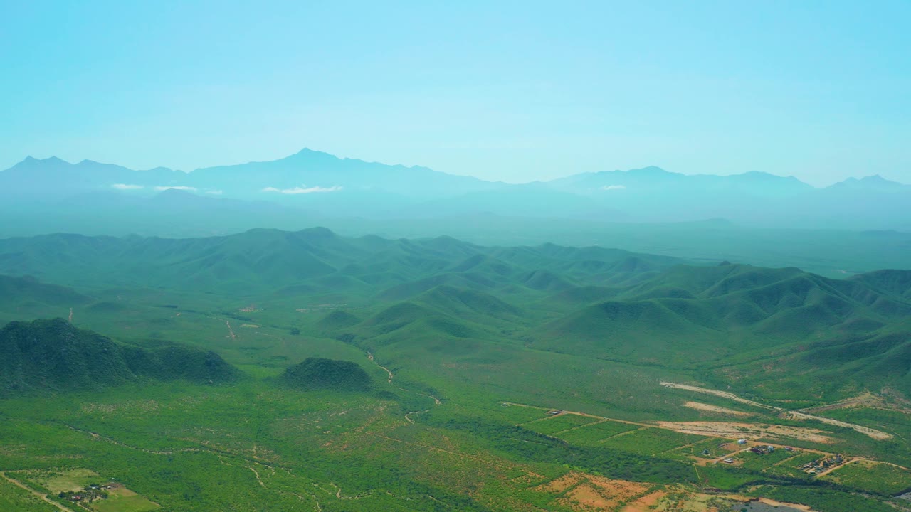 High aerial frame from Cerritos Beach, Baja California Sur, with rounded green hills across the valley, light-colored dirt roads below, distant ridgelines, and no visible people.