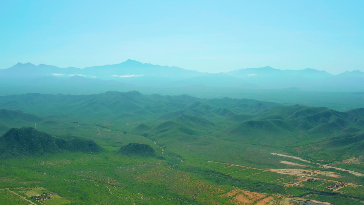 Wide drone-like view over Cerritos Beach inland terrain with green hills in front, pale tracks crossing the valley floor, hazy mountains in back, and no visible people.