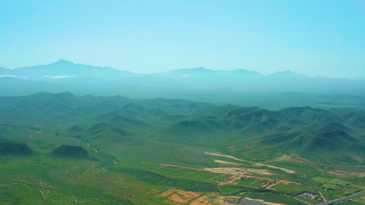 Aerial view one second into the Cerritos Beach valley clip, with rounded green hills centered below a hazy mountain ridge and no visible people.