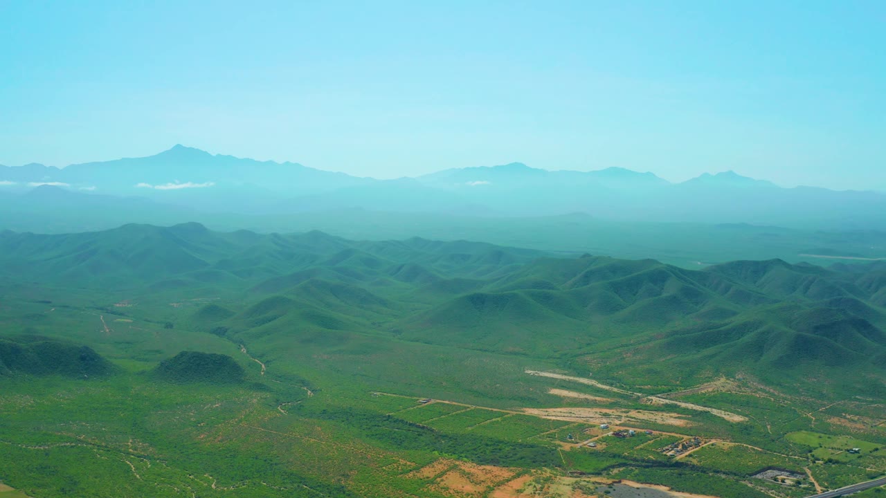 Aerial frame over the Baja California Sur valley with layered blue mountains, low green hills, pale tracks, and no visible people.