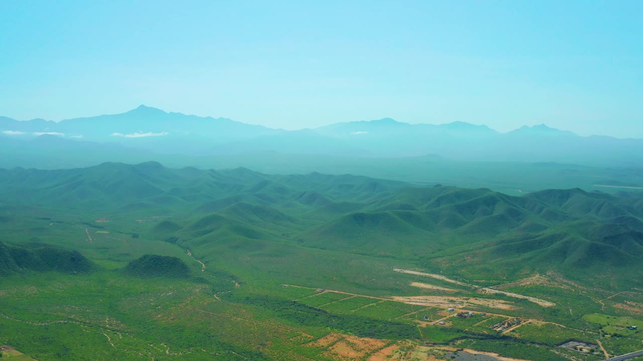 Mid-clip aerial view of Cerritos Beach inland terrain, showing green fields, rounded hills, faint roads, mountain silhouettes, and no visible people.