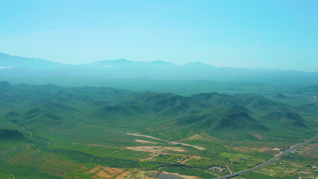 Opening aerial frame over Cerritos Beach, Baja California Sur, showing green rolling hills, a road in the lower foreground, hazy mountains on the horizon, a small white cloud at upper left, and no visible people.