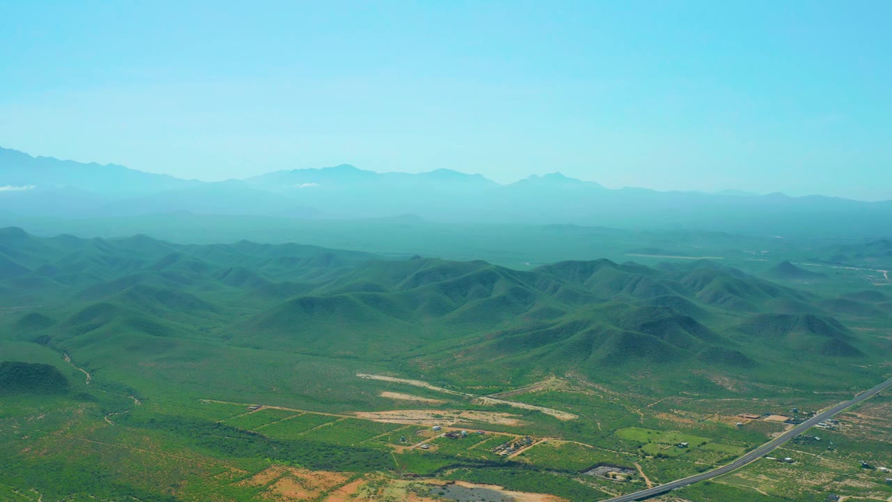 Aerial frame from Cerritos Beach, Baja California Sur, with the paved road angled through the lower right, green hill forms across the middle distance, pale dirt clearings, hazy mountains, blue sky, and no visible people.