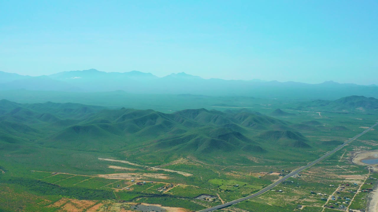 Wide aerial frame over Cerritos Beach, Baja California Sur countryside with green rolling hills, a paved road in the valley, hazy mountains on the horizon, and no visible people.