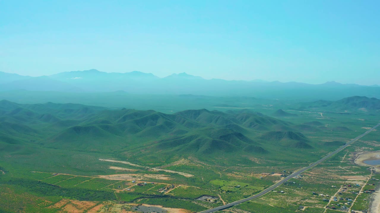 Aerial view in Cerritos Beach, Baja California Sur showing the valley road more centrally, with green hills in the midground, pale field patches below, and no visible people.
