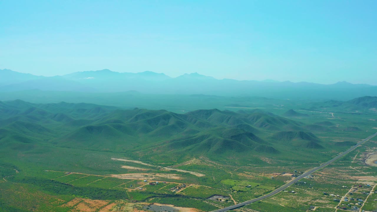Cerritos Beach, Baja California Sur aerial still with a broad blue sky, soft haze over distant mountain ridges, green hills in the midground, and no visible people.