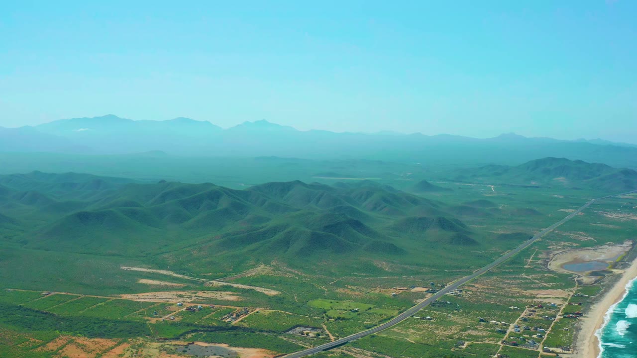 High aerial frame over Cerritos Beach, Baja California Sur, showing a coastal highway beside the sandy beach, green inland hills, distant mountain ridges, blue sky, and no visible people.