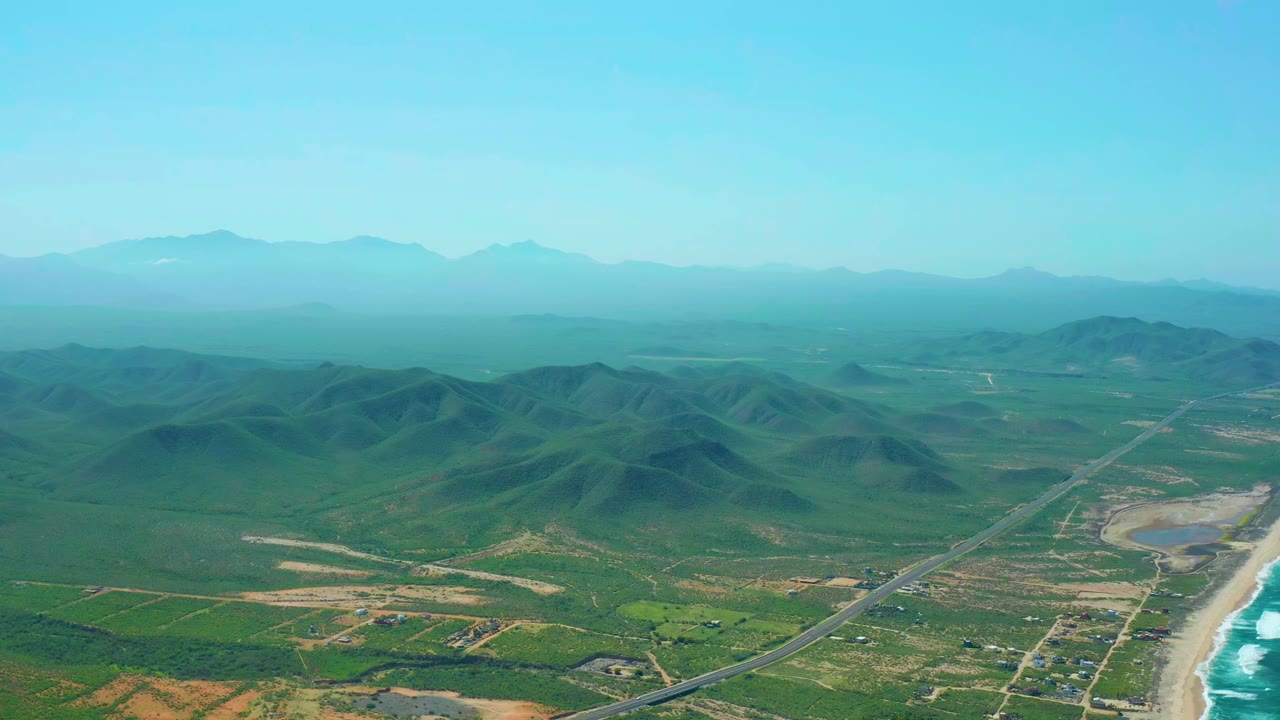 Aerial view in Cerritos Beach with the highway cutting through green terrain toward the beach, a bright surf line at right, hazy mountain background, and no visible people.