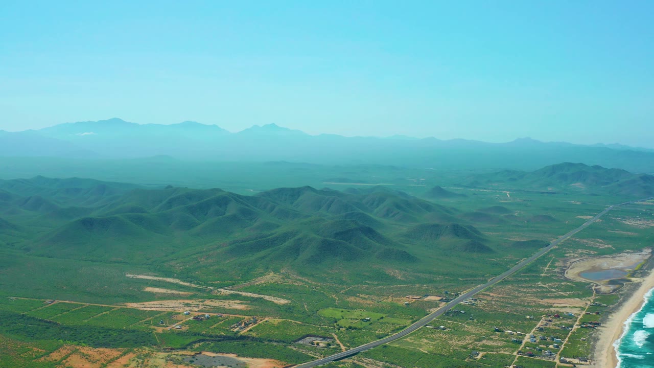 Cerritos Beach aerial frame with the road angled through the lower landscape, low hills centered in the frame, ocean surf along the right edge, distant ridges, and no visible people.