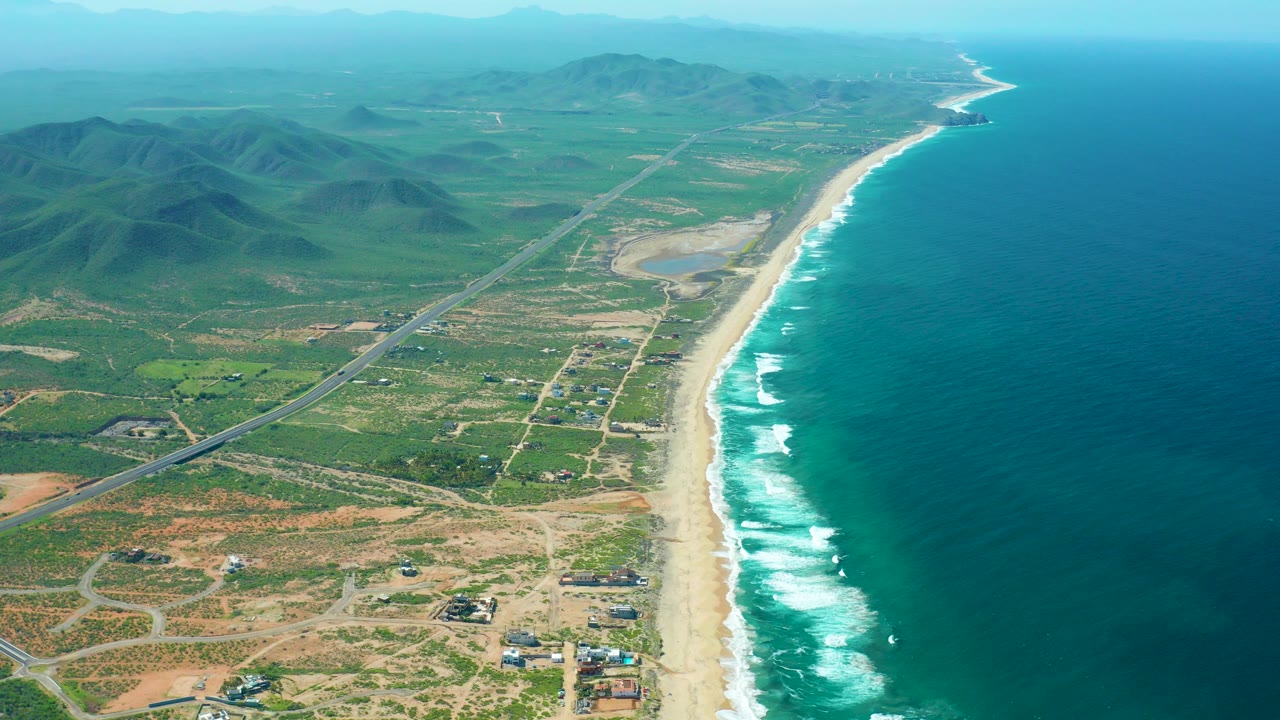 Aerial view of beach, ocean, coastal road, green land, and mountains.