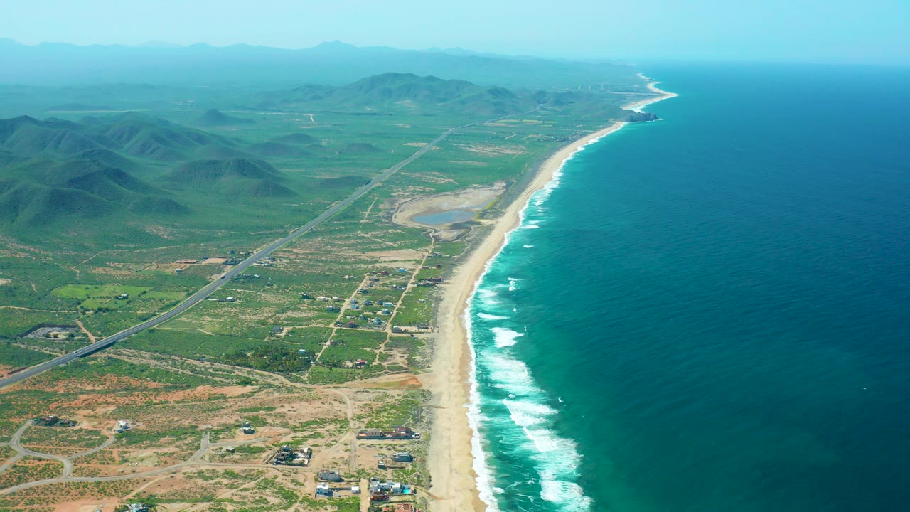 Opening aerial view of Cerritos Beach in Baja California Sur with a sandy shoreline, blue-green ocean, white surf, inland road, green hills, hazy sky, and no visible people.