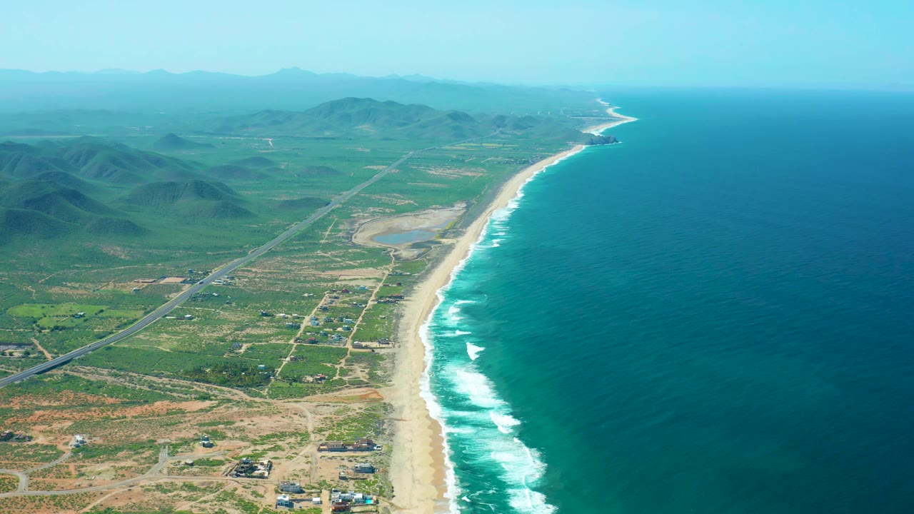 High aerial frame over Cerritos Beach with the shoreline running diagonally, surf bands along the sand, inland roads, low hills, distant hazy mountains, and no visible people.