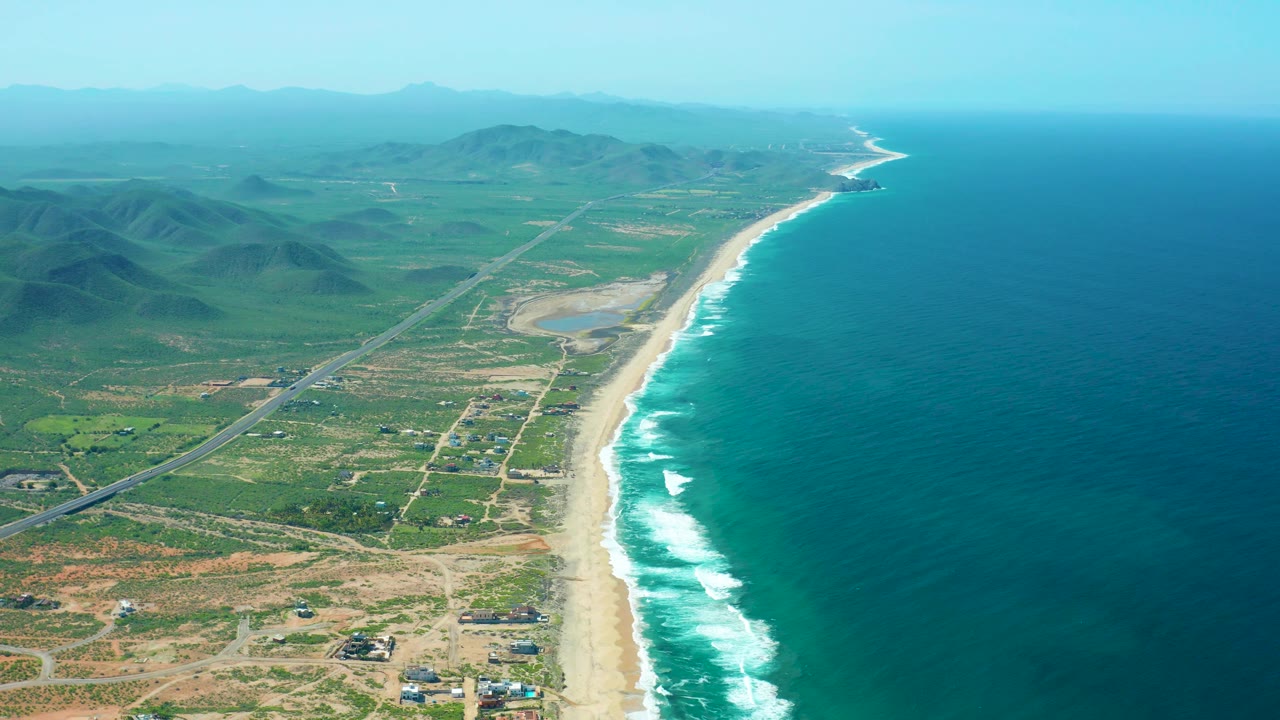 Drone view of Cerritos Beach with deep blue ocean to the right, pale surf along the coast, patchy green inland terrain, roads, a small lagoon-like water area, hazy hills, and no visible people.
