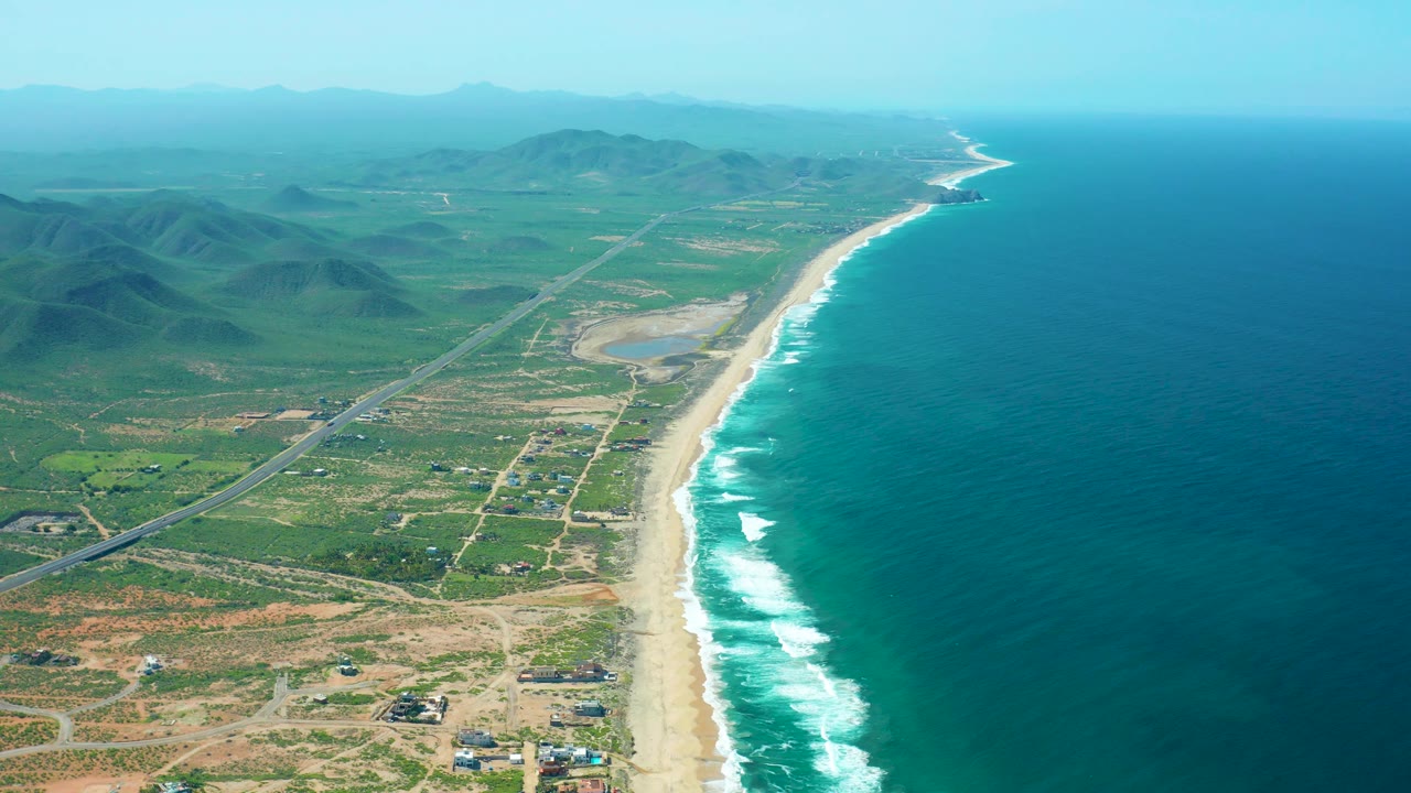 Aerial coastal landscape with waves, beach, roads, and mountains in haze.