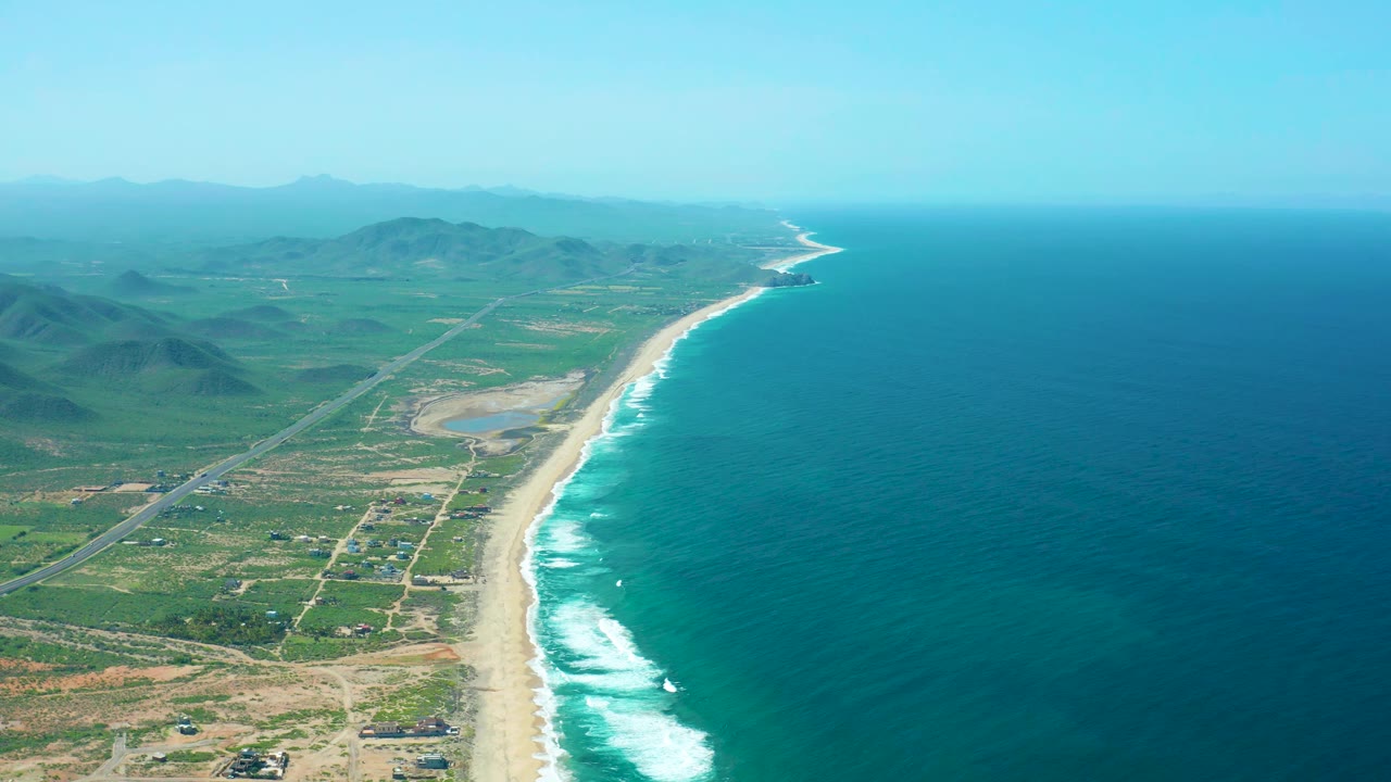 Wide aerial coastline with surf, road, fields, hills, and ocean.