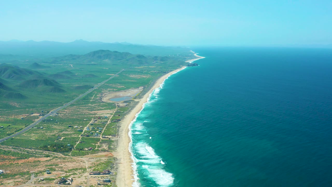 Cerritos Beach aerial with more open ocean filling the right side, a pale sandy beach and bright surf band below, rural fields and roads inland, hazy mountains on the horizon, and no visible people.