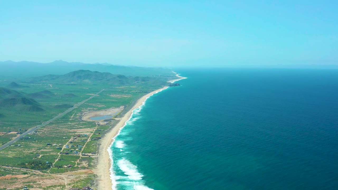 Aerial view along Cerritos Beach with white surf marking the shore, inland roads crossing green terrain, hazy mountain ridges in the distance, and no visible people.