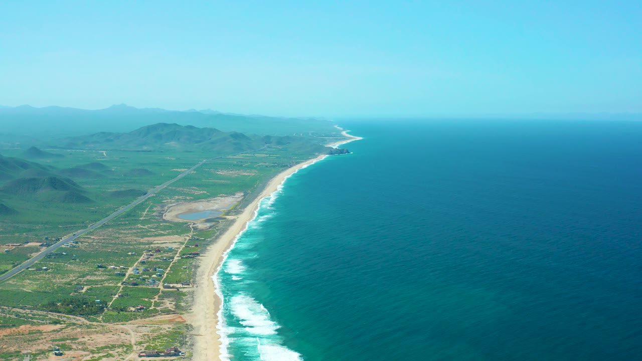 Mid-clip aerial view of Cerritos Beach showing rolling surf along the sand, a green coastal plain with roads and scattered structures, hazy mountains, and no visible people.