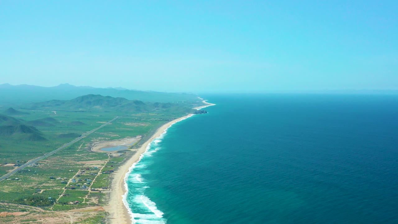 Opening aerial frame over Cerritos Beach, Baja California Sur, with blue ocean to the right, white surf along the sand, green inland fields, roads, distant mountains, and no visible people.