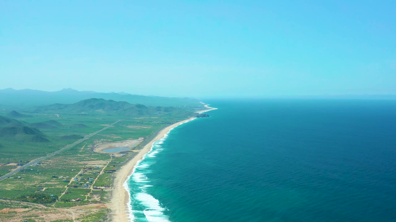 Aerial view over Cerritos Beach with the white wave line slightly farther along the sand, ocean occupying the right side, green and tan land with road grids on the left, hazy mountains beyond, and no visible people.