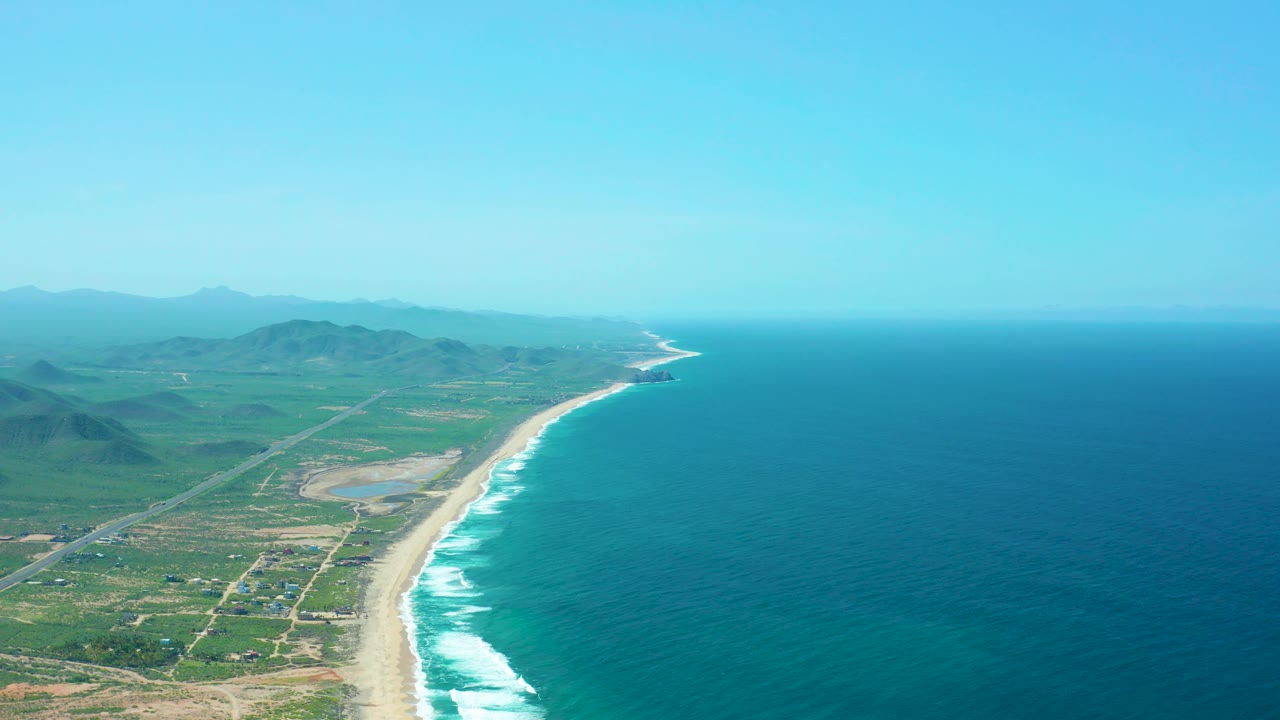 Mid-clip aerial frame looking along Cerritos Beach, with a long sand strip and active white surf between the blue ocean and green-brown coastal land, roads and small structures inland, distant mountains, and no visible people.