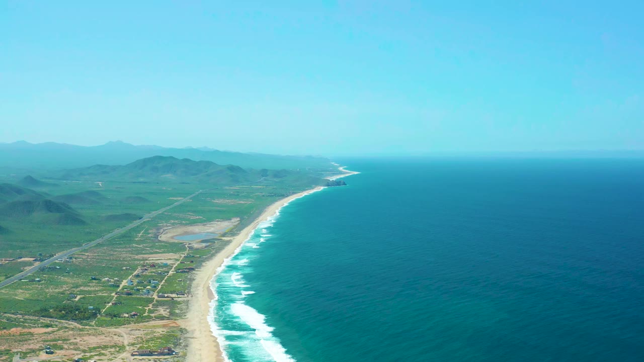 Aerial view over Cerritos Beach showing the coastal road and small vehicle area inland from the sand, with waves breaking along the shore and no visible people.