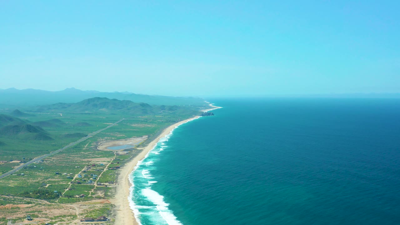 Cerritos Beach aerial frame with the ocean occupying the right half, the sandy surf line curving into the distance, rural roads inland, green hills beyond, and no visible people.