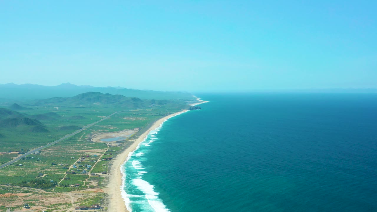 Mid-clip aerial over Cerritos Beach with white wave bands along the sand, a coastal road crossing the green inland area, distant hills, hazy blue sky, and no visible people.