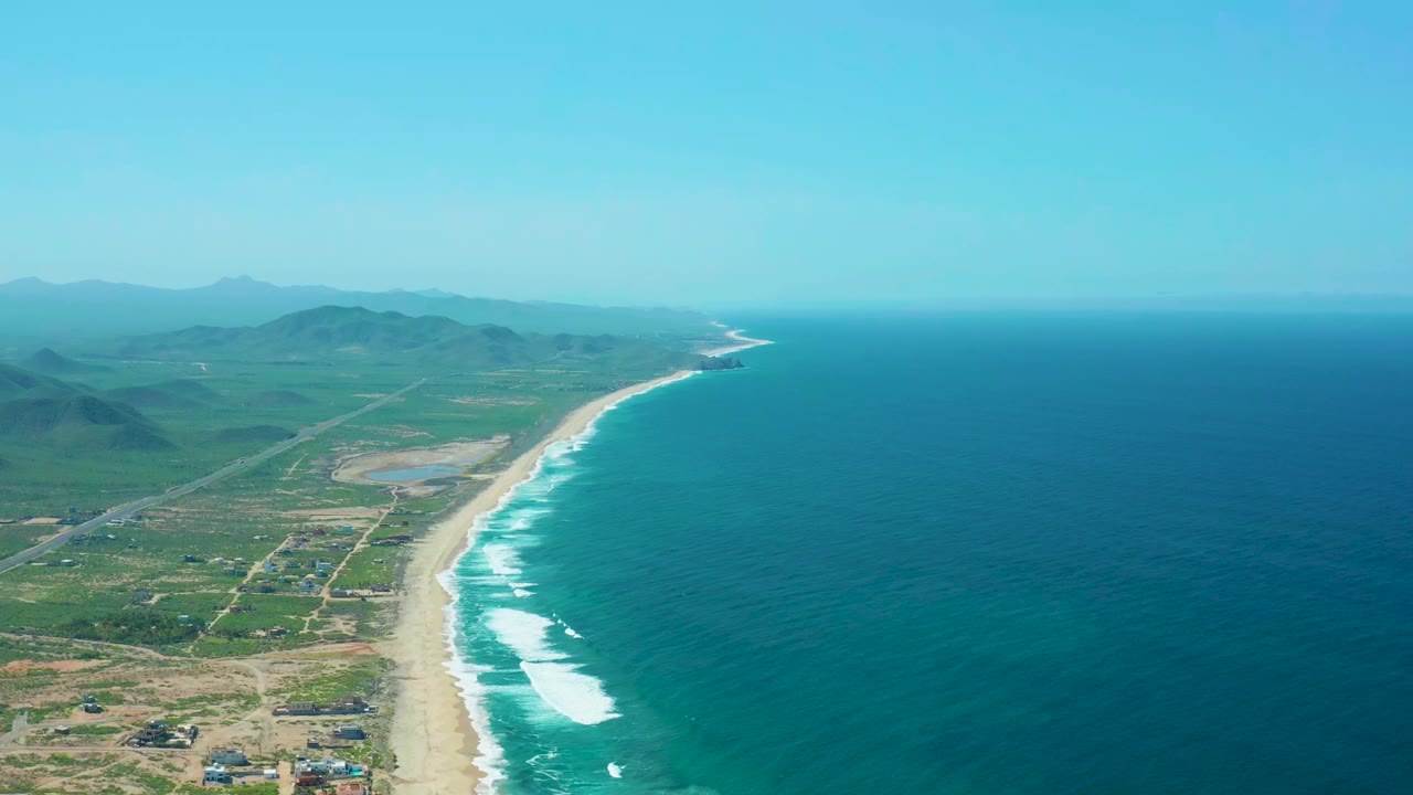 Cerritos Beach aerial view at the start of the clip, with blue ocean on the right, a sandy shoreline, green land and roads on the left, distant hills, and no visible people.