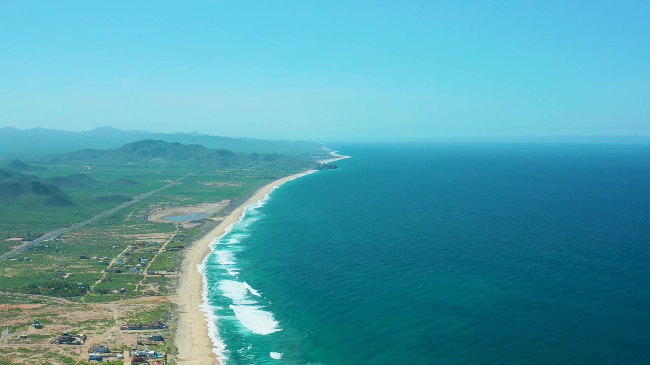 Wide aerial coastline with beach, ocean, roads, and distant mountains.