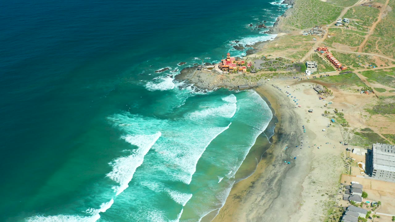 Opening aerial frame over Cerritos Beach, Baja California Sur, with deep blue water at left, white surf reaching the sand, a rocky point above the beach, scattered buildings, and no visible people.