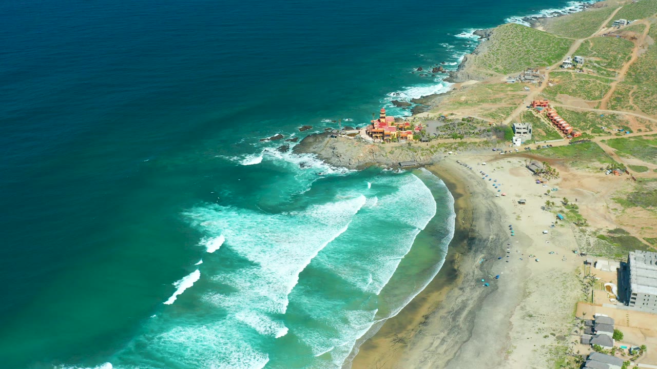 Aerial frame over Cerritos Beach, Baja California Sur, with turquoise water, white surf, sandy shore, rocky headland, coastal buildings, and no visible people.