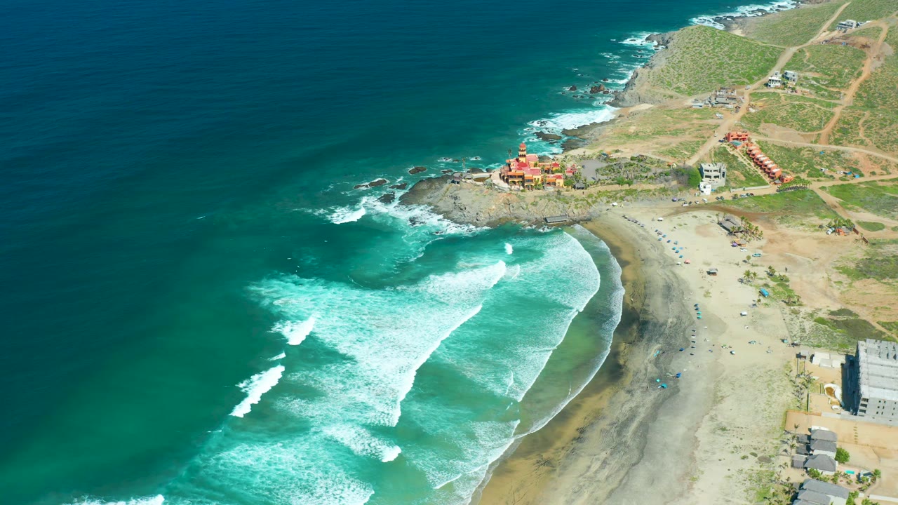 Aerial view of Cerritos Beach with whitewater near the shore, a rocky point above the beach, low buildings inland, and no visible people.