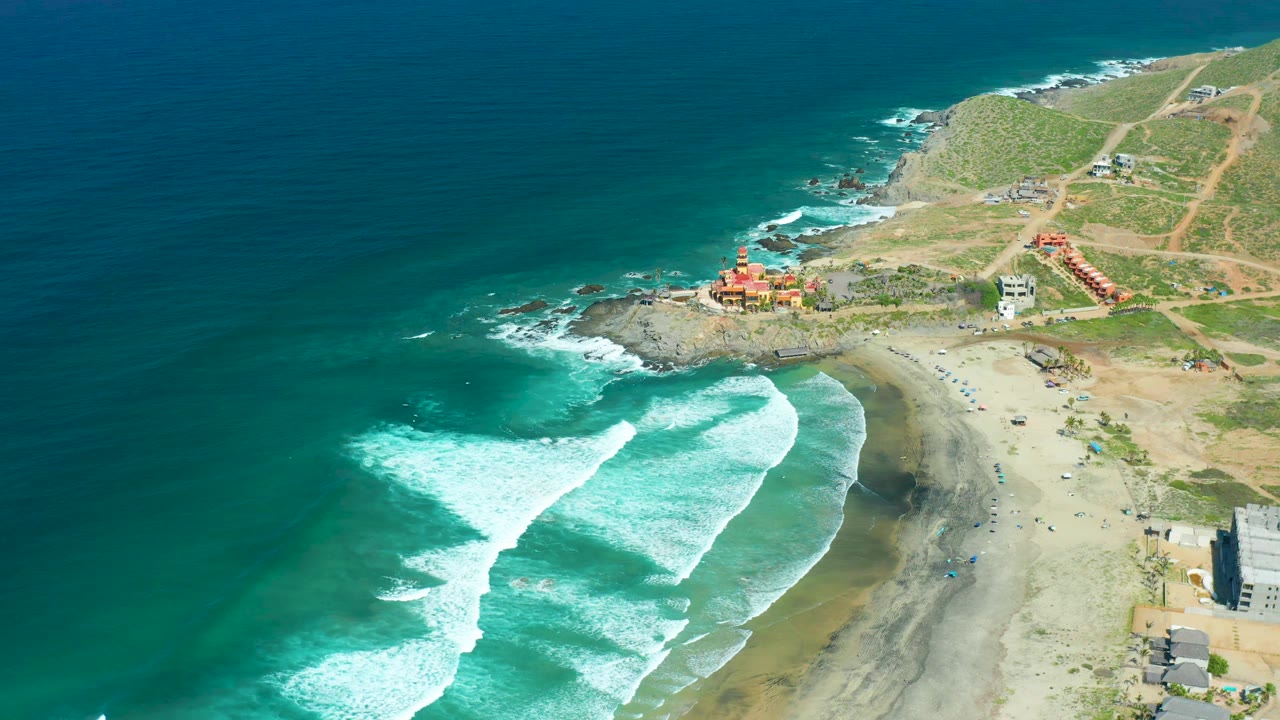 Cerritos Beach aerial view with white surf spreading wider across turquoise water, sandy beach below, hillside roads and small coastal structures in the background, and no visible people.
